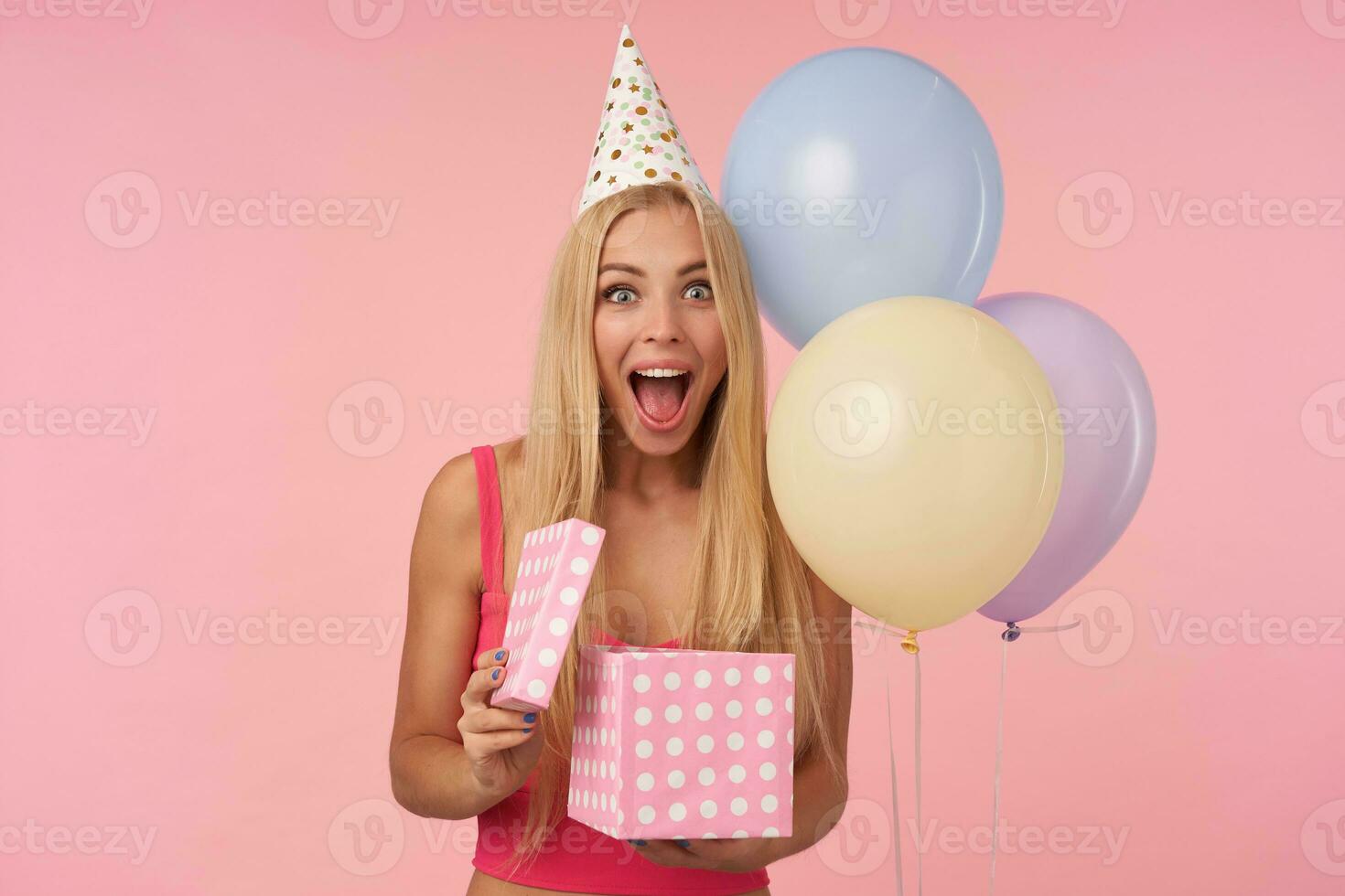 überglücklich jung blond weiblich im Rosa oben und Urlaub Hut Sein aufgeregt und überrascht zu erhalten Geburtstag die Geschenke, Stehen Über Rosa Hintergrund mit Bündel von Helium Luftballons foto