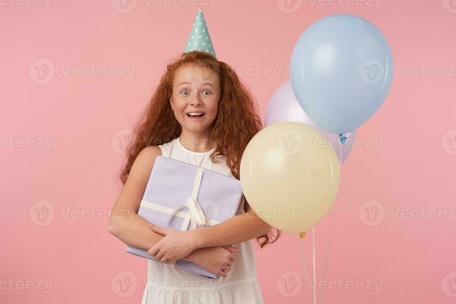 freudig weiblich Kind mit lange foxy Haar Sein aufgeregt und überrascht zu erhalten Geburtstag gegenwärtig, Stehen Über Rosa Hintergrund mit Geschenk im Hände und suchen im Kamera mit breit glücklich Lächeln foto