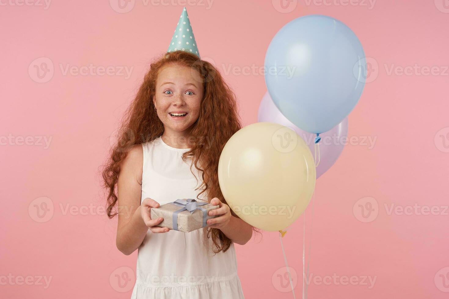 Studio Schuss von froh Rothaarige wenig Mädchen mit lange lockig Haar halten Geschenk verpackt Box Sein aufgeregt und überrascht zu erhalten Geburtstag gegenwärtig, Stehen Über Rosa Hintergrund im festlich Kleider foto