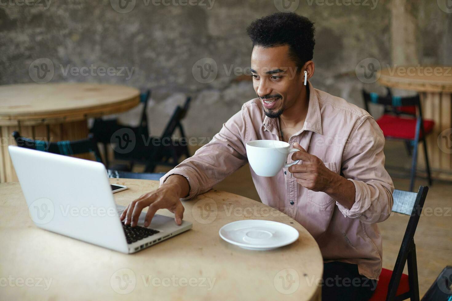 heiter jung bärtig Mann mit dunkel Haut Arbeiten im Kaffee Haus mit modern Laptop mit Öffentlichkeit W-lan, lesen Mail und haben gut Tag, behalten Tasse von Tee im seine Hand und lächelnd foto