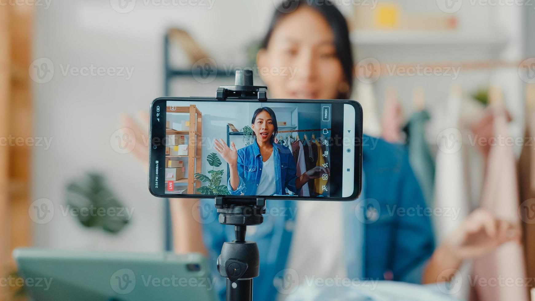 junge asiatische Modedesignerin mit Handy, die eine Bestellung entgegennimmt und Kleidung zeigt, die Video-Live-Streaming online im Geschäft aufzeichnet. Kleinunternehmer, Online-Markt-Bereitstellungskonzept. foto