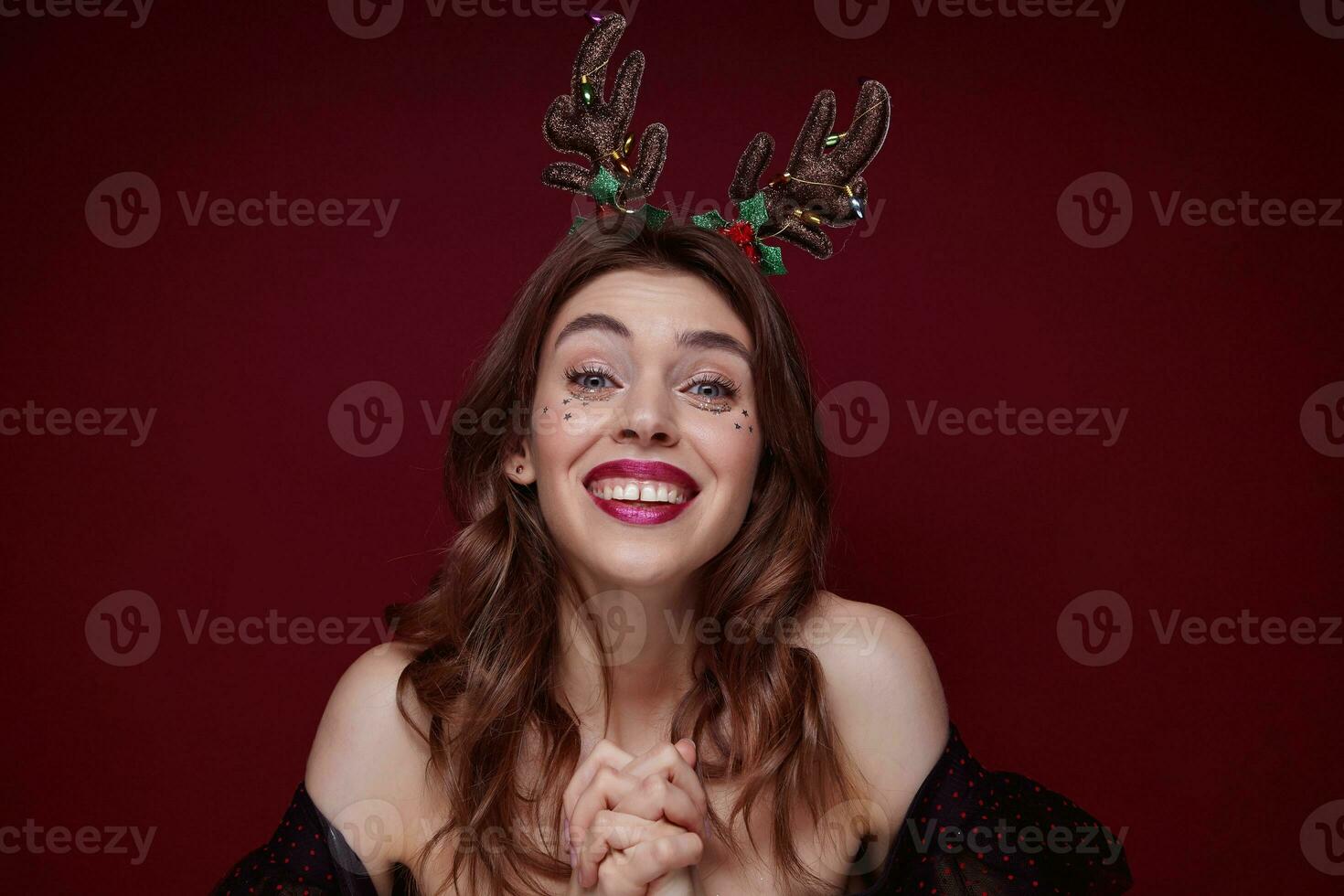 Innen- Schuss von jung froh schön Brünette mit wellig Frisur genießen Weihnachten Thema Party Maskerade und lächelnd glücklich zu Kamera, gekleidet im elegant Kleider Über Rotwein Hintergrund foto