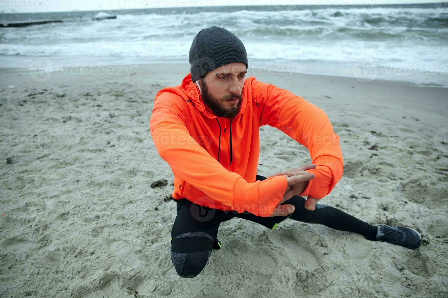 Bild von ernst suchen jung bärtig Sportler gekleidet im warm sportlich Kleider tun Morgen Übungen durch das Meer, Dehnen nach Laufen Sitzung. Fitness und Sport Konzept foto