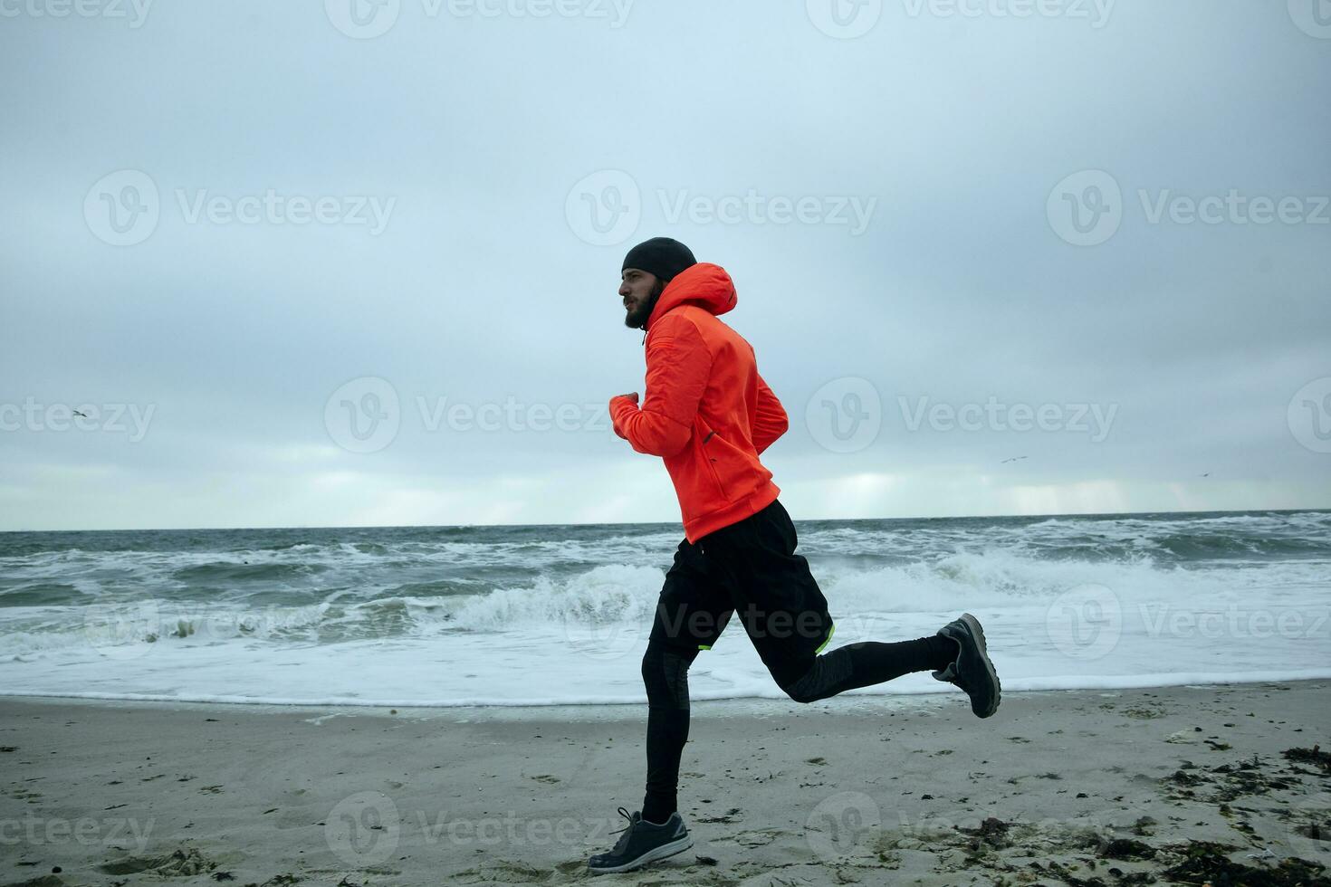 Schuss von sportlich jung bärtig Mann im schwarz Deckel, sportlich Kleider und warm Orange Mantel Laufen auf Küste von Meer auf grau stürmisch Wetter. Sport und gesund Lebensstil Konzept foto