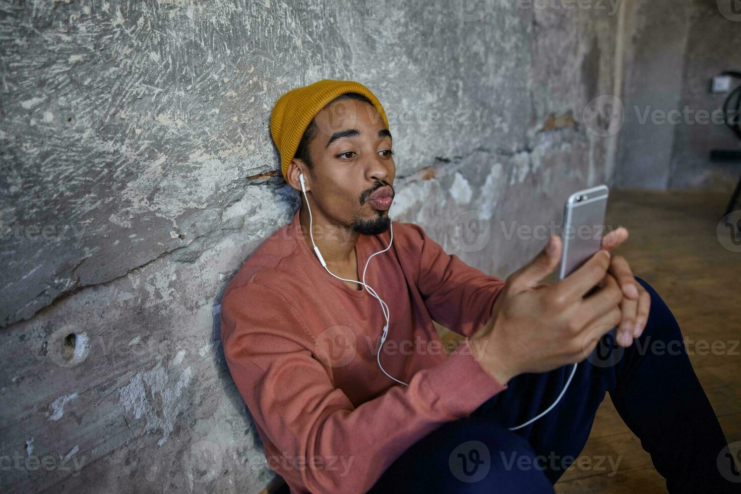 Innen- Foto von ziemlich bärtig dunkel gehäutet Kerl im Rosa Pullover, Blau Hose, Hose und Senf Deckel gelehnt auf Beton Wand, Herstellung Foto von selbst mit Handy, Mobiltelefon Telefon und falten Lippen im Luft Kuss