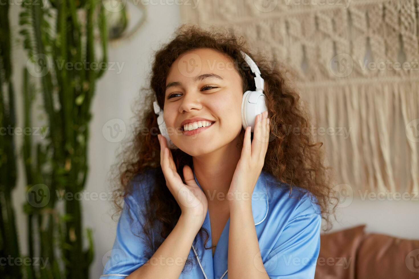 Porträt von lockig jung Mulatte Dame, breit lächelt mit geschlossen Augen, Hören Liebling Lied im Kopfhörer, genießen das Musik- und sonntags Morgen, sieht aus glücklich und froh. foto