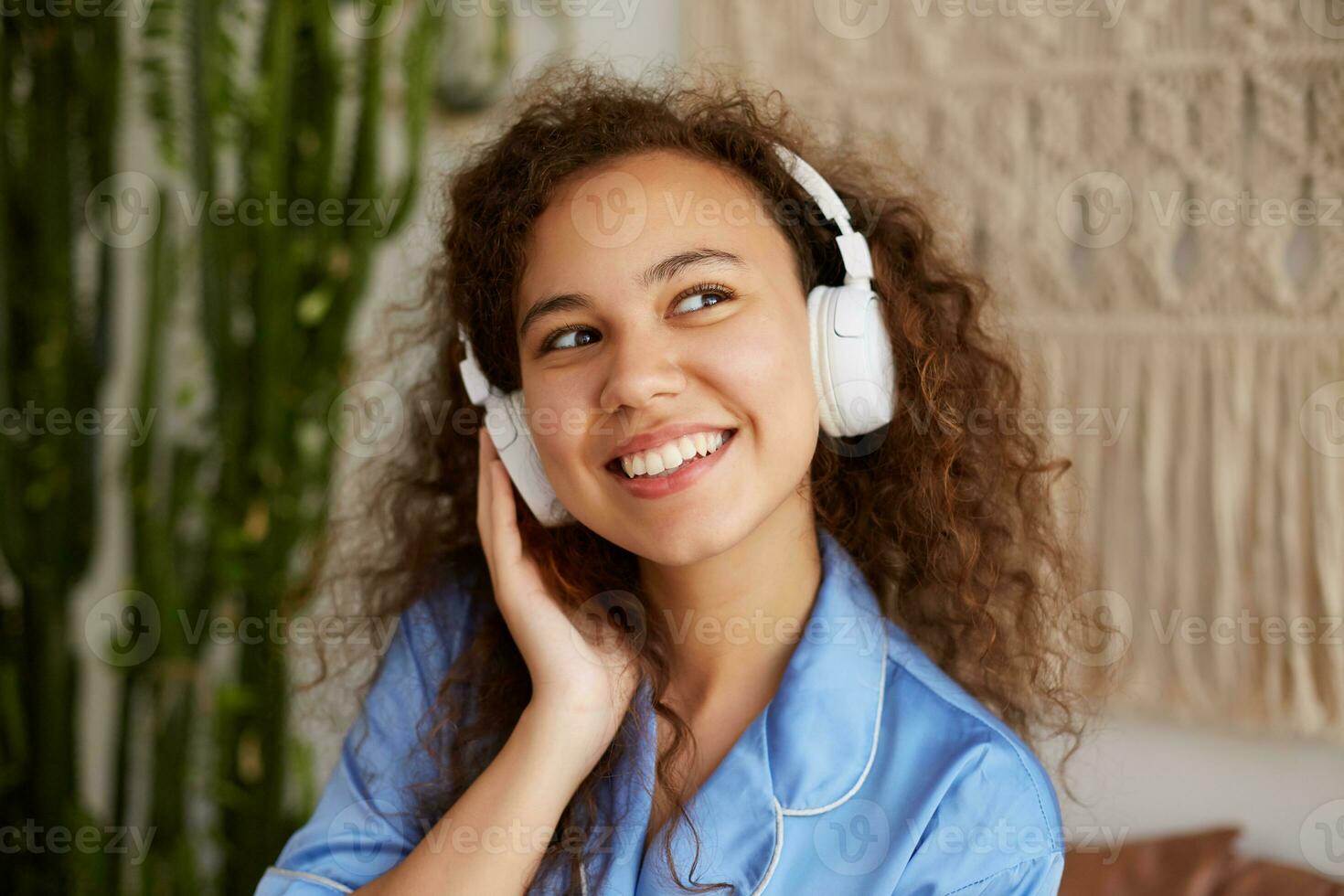 glücklich lockig jung Mulatte jung Frau, breit lächelt und Hören Liebling Lied im Kopfhörer, genießen das Musik- und sonntags Morgen, sieht aus glücklich und froh. foto