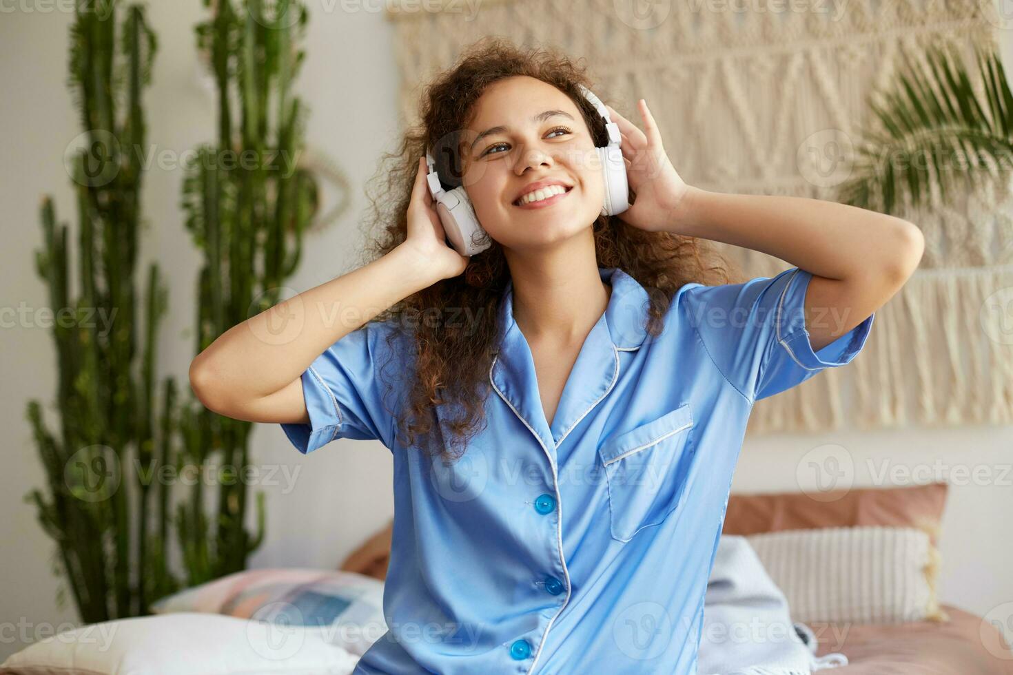 Foto von lockig jung nett afrikanisch amerikanisch Dame, breit lächelt mit geschlossen Augen, Hören Liebling Musik- im Kopfhörer, hält Kopfhörer, genießen das sonntags Morgen, sieht aus glücklich und froh.