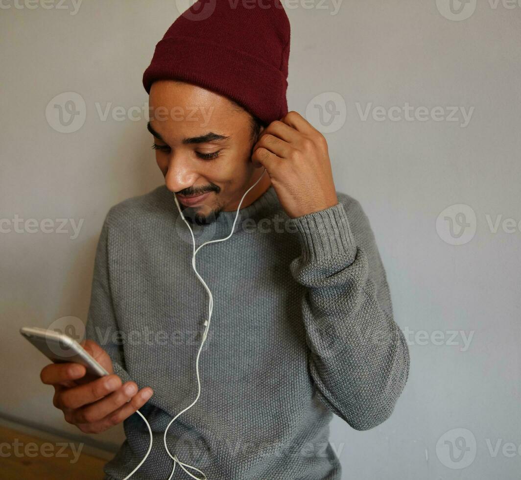 Nahansicht von charmant jung dunkel gehäutet männlich mit Bart Einfügen Hörmuschel in seine Ohr und suchen positiv auf Bildschirm von seine Telefon, tragen beiläufig Kleider foto