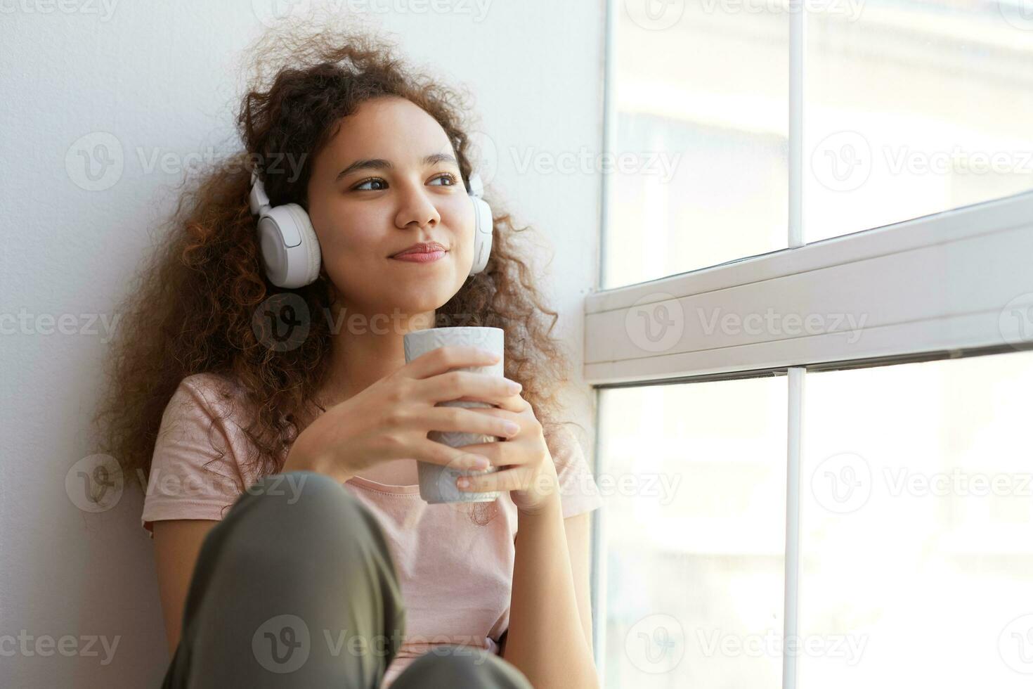 träumend jung afrikanisch amerikanisch Frau mit lockig Haar, Hören Favorit Lied beim Kopfhörer und Trinken Tee, sieht aus beim das Fenster und genießen sonnig Tag beim heim. foto