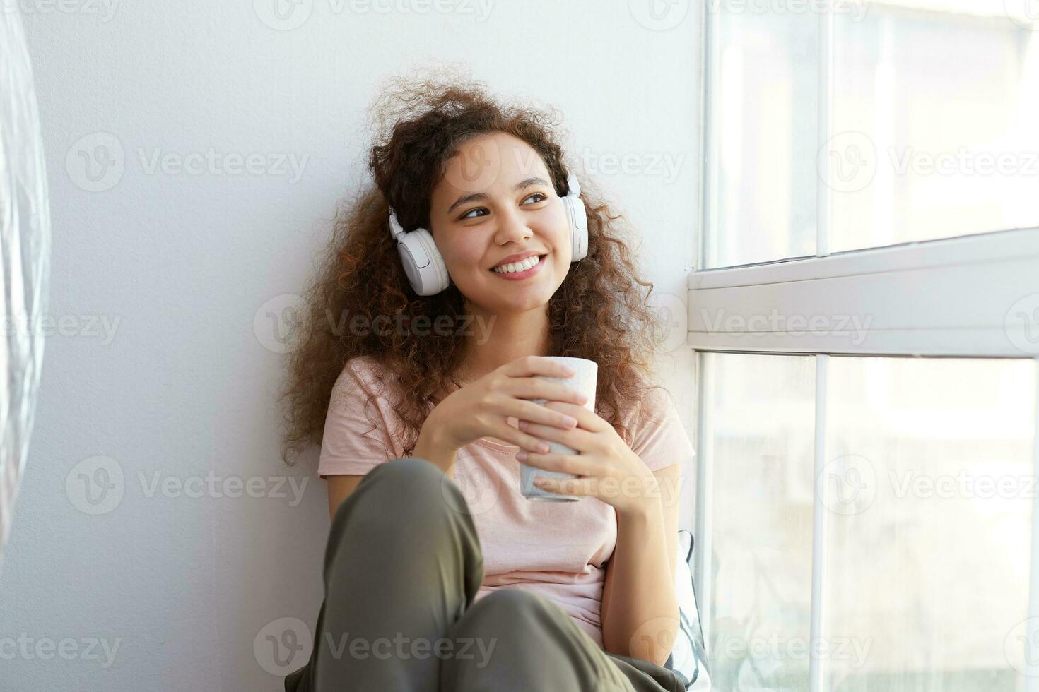 glücklich jung afrikanisch amerikanisch Frau Sitzung durch das Fenster, Hören Favorit Lied beim Kopfhörer und Trinken Tee, sieht aus beim das Fenster und genießen das sonnig Tag beim heim. foto