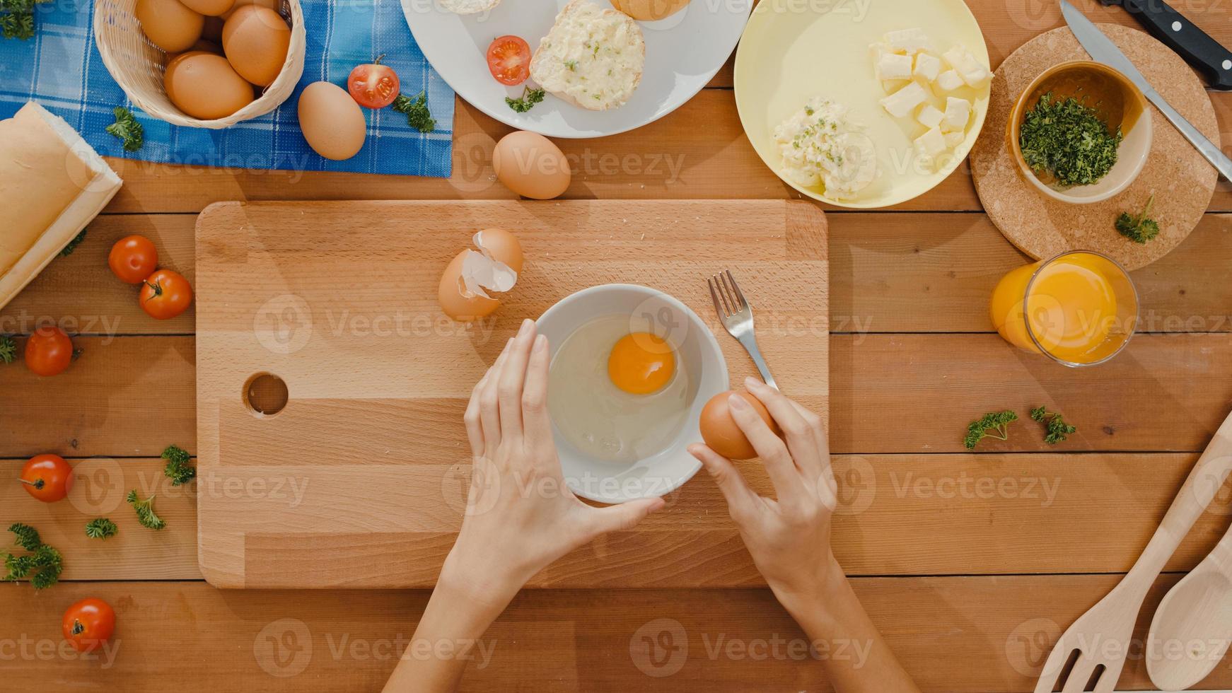 Hände der jungen asiatischen Köchin knacken Ei in Keramikschale kochen Omelett mit Gemüse auf Holzbrett auf dem Küchentisch im Haus. Lifestyle gesundes Essen und traditionelles Bäckereikonzept. Ansicht von oben geschossen. foto