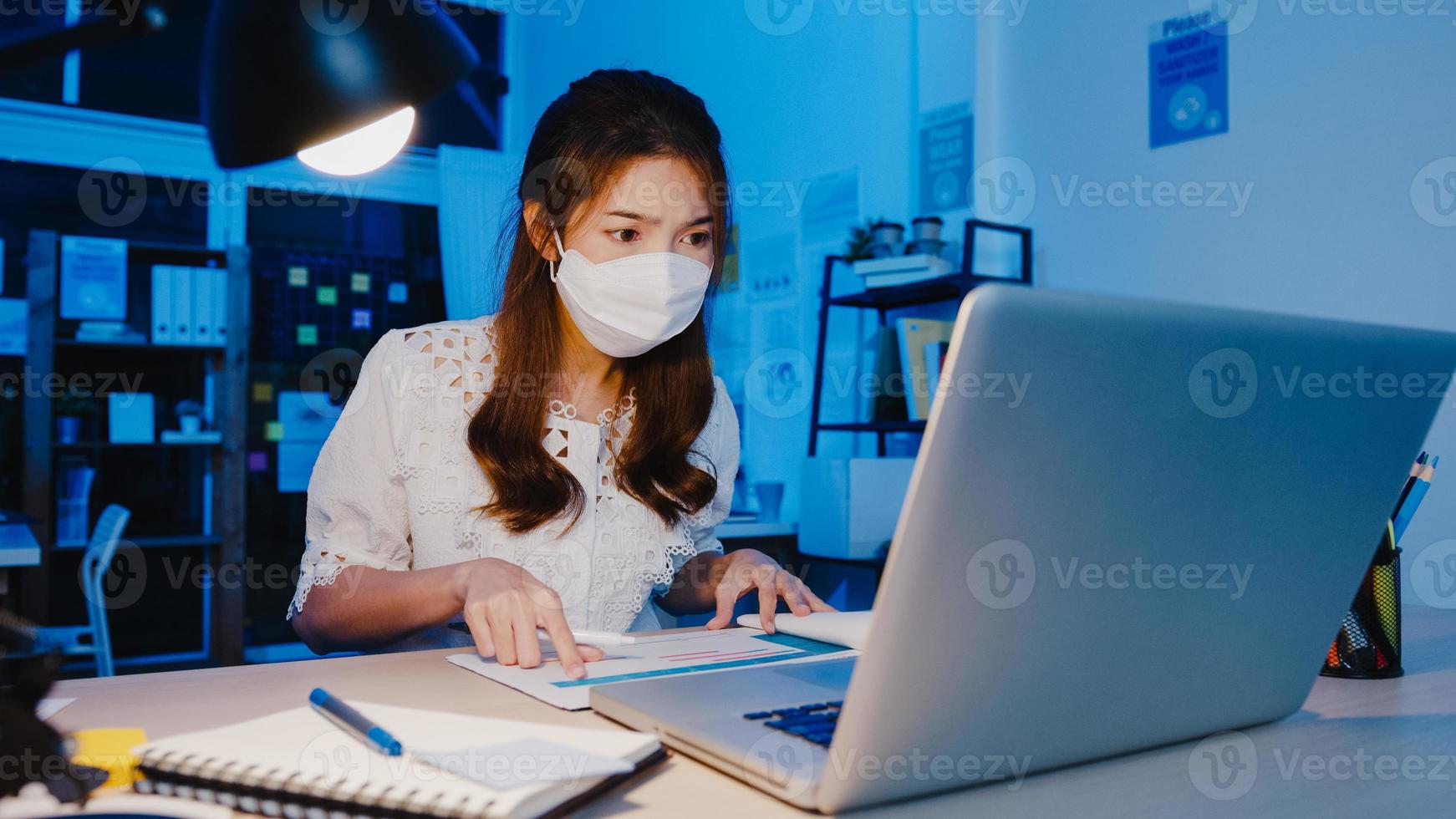 Fröhliche Geschäftsfrau in Asien, die eine medizinische Gesichtsmaske für soziale Distanzierung in einer neuen normalen Situation zur Virenprävention trägt, während sie in der Büronacht den Laptop bei der Arbeit verwendet. Leben und Arbeiten nach dem Coronavirus. foto