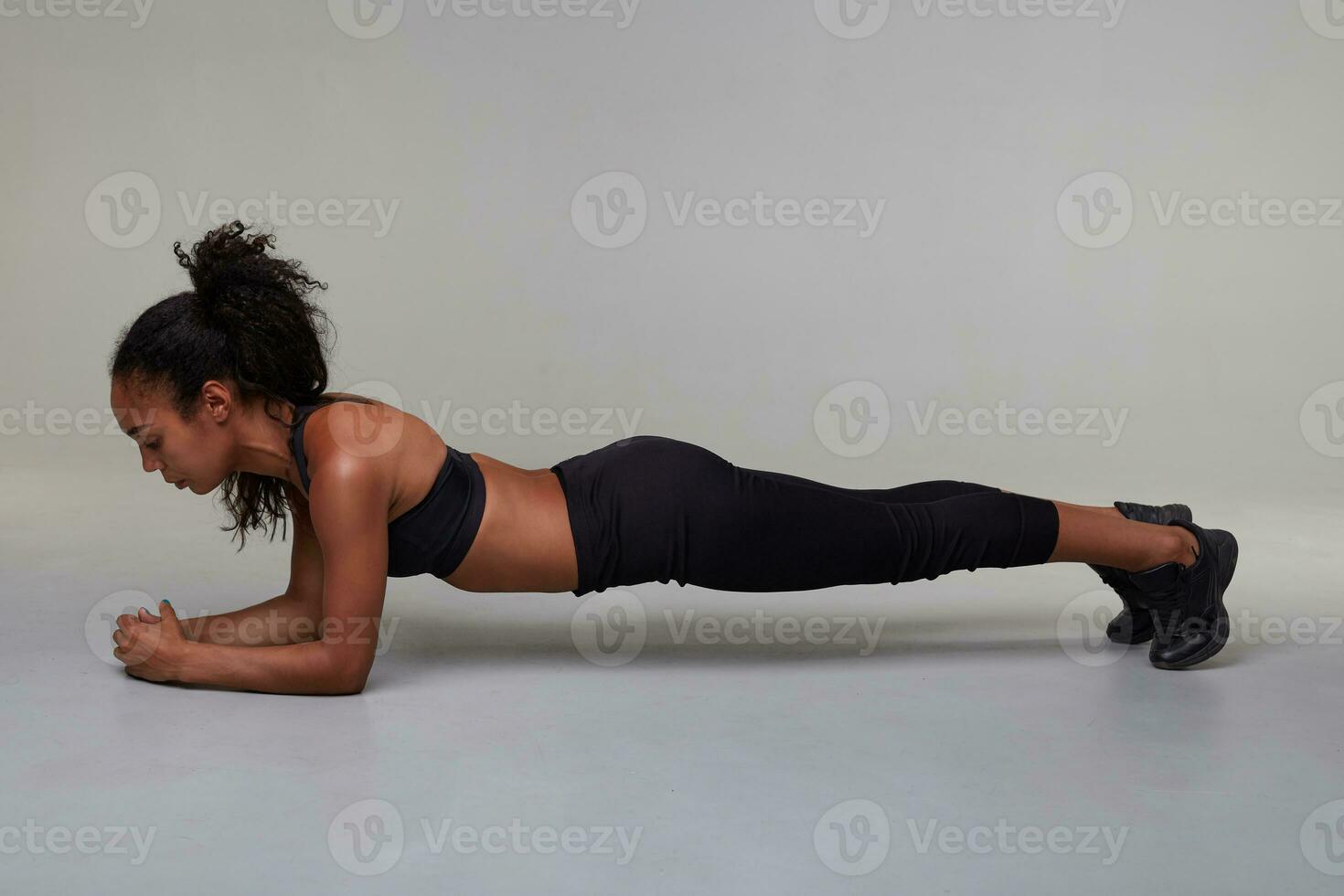 in voller Länge Foto von jung sportlich dunkel gehäutet lockig weiblich mit gut Körper tun Planke Übung drinnen, Stehen Über grau Hintergrund im schwarz oben und Leggings. Sport und Wellness Konzept