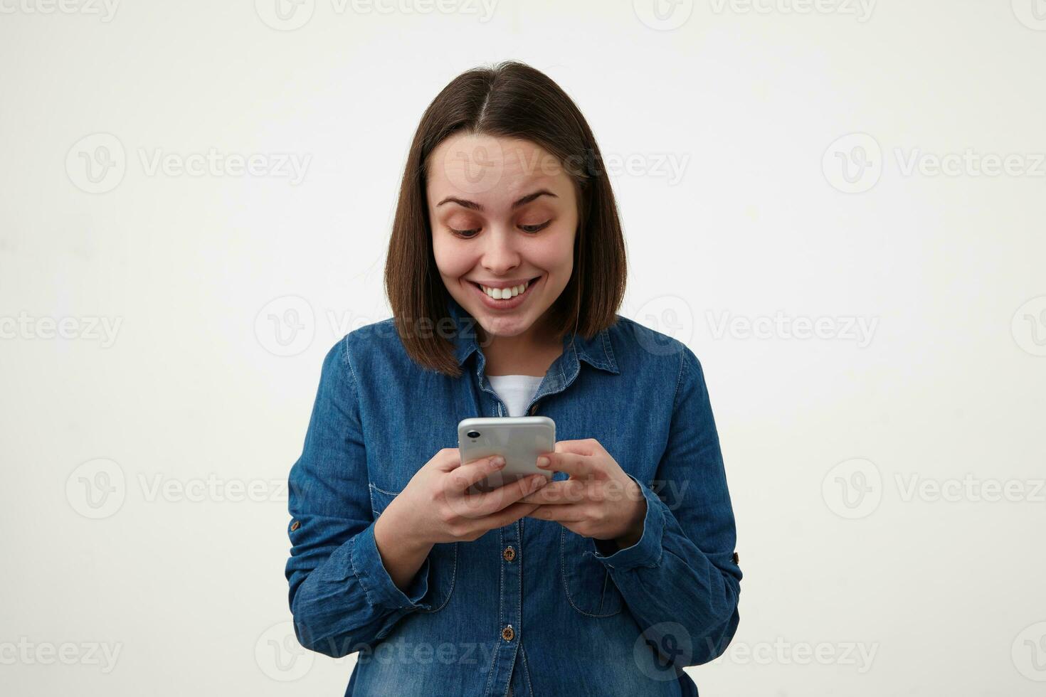froh jung ziemlich kurz behaart Brünette Dame mit natürlich bilden behalten ihr Handy, Mobiltelefon Telefon im angehoben Hände und lächelnd glücklich während suchen beim Bildschirm, posieren Über Weiß Hintergrund foto