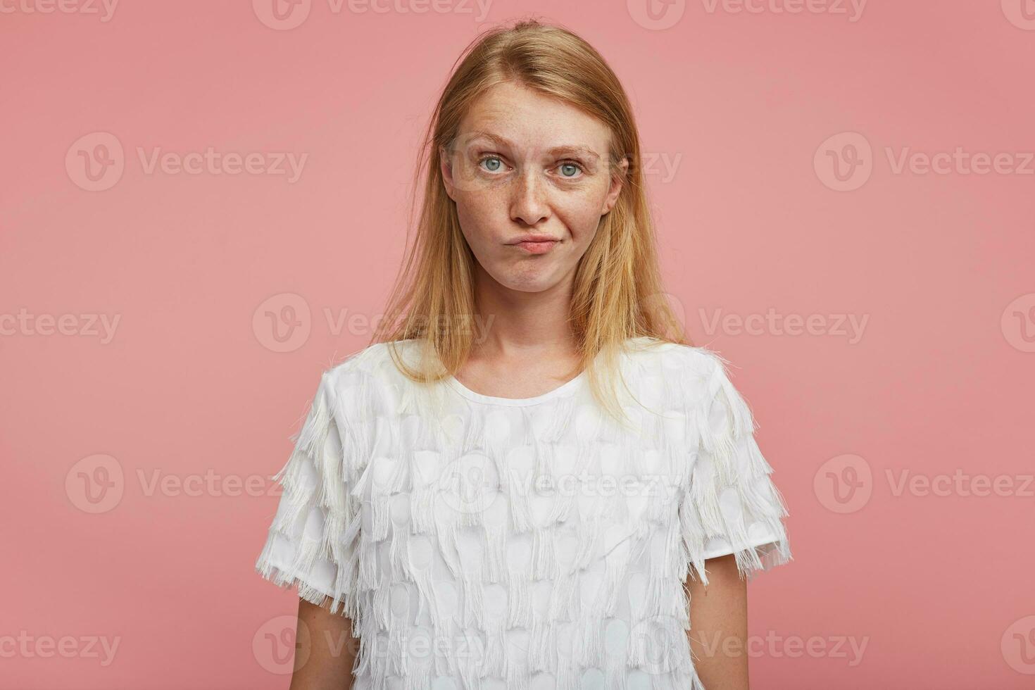 unzufrieden jung ziemlich Rothaarige weiblich mit Grün Grau Augen Verdrehen ihr Mund während suchen verwirrt zu Kamera, Stehen Über Rosa Hintergrund mit Hände Nieder foto