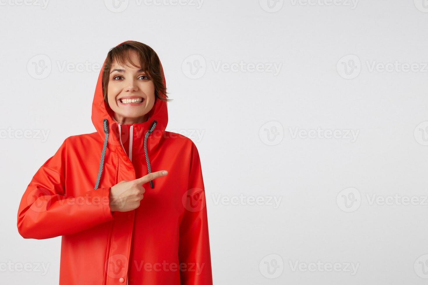 Porträt von ung glücklich breit lächelnd süß kurz behaart Frau gekleidet im rot Regen Mantel, will zu zeichnen Ihre Aufmerksamkeit, Punkte Finger zu das Kopieren Raum. Stehen Über Weiß Hintergrund. foto