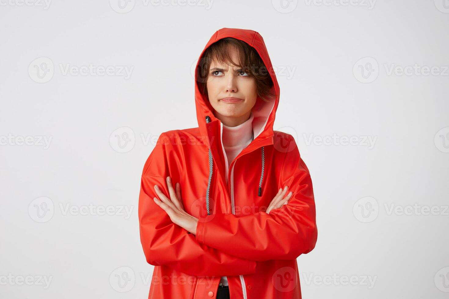 jung unzufrieden mit regnerisch Wetter süß kurz behaart Dame gekleidet im rot Regen Mantel, mit ein Kapuze auf seine Kopf, suchen Weg mit traurig Ausdruck, Stehen Über Weiß Hintergrund. foto