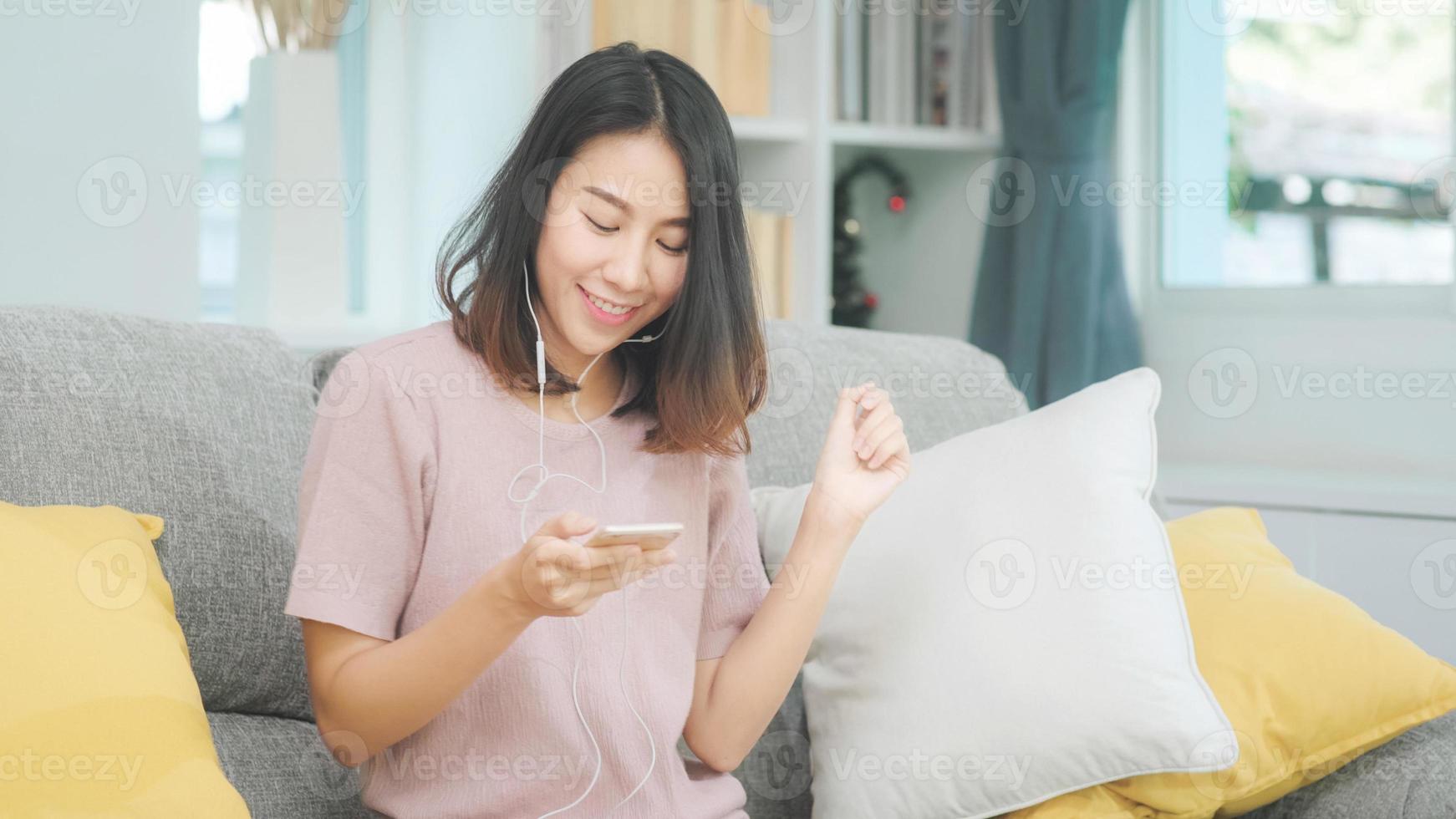 asiatische Frau, die Musik hört und Smartphone verwendet, Frau mit Entspannungszeit auf dem Sofa im Wohnzimmer zu Hause liegend. glückliche weibliche hörende musik mit kopfhörerkonzept. foto