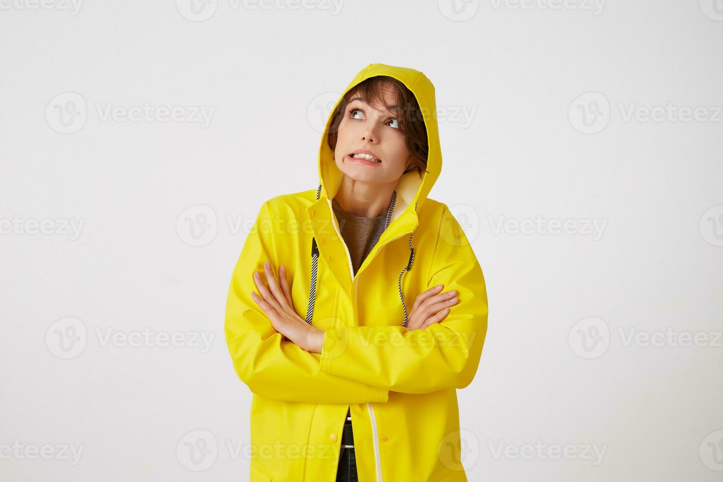 Porträt von jung angeekelt süß kurz behaart Mädchen trägt im Gelb Regen Mantel, sieht aus oben mit unzufrieden Ausdrücke, versteckt unter ein Regen Haube, steht Über Weiß Mauer mit gekreuzt Waffen. foto