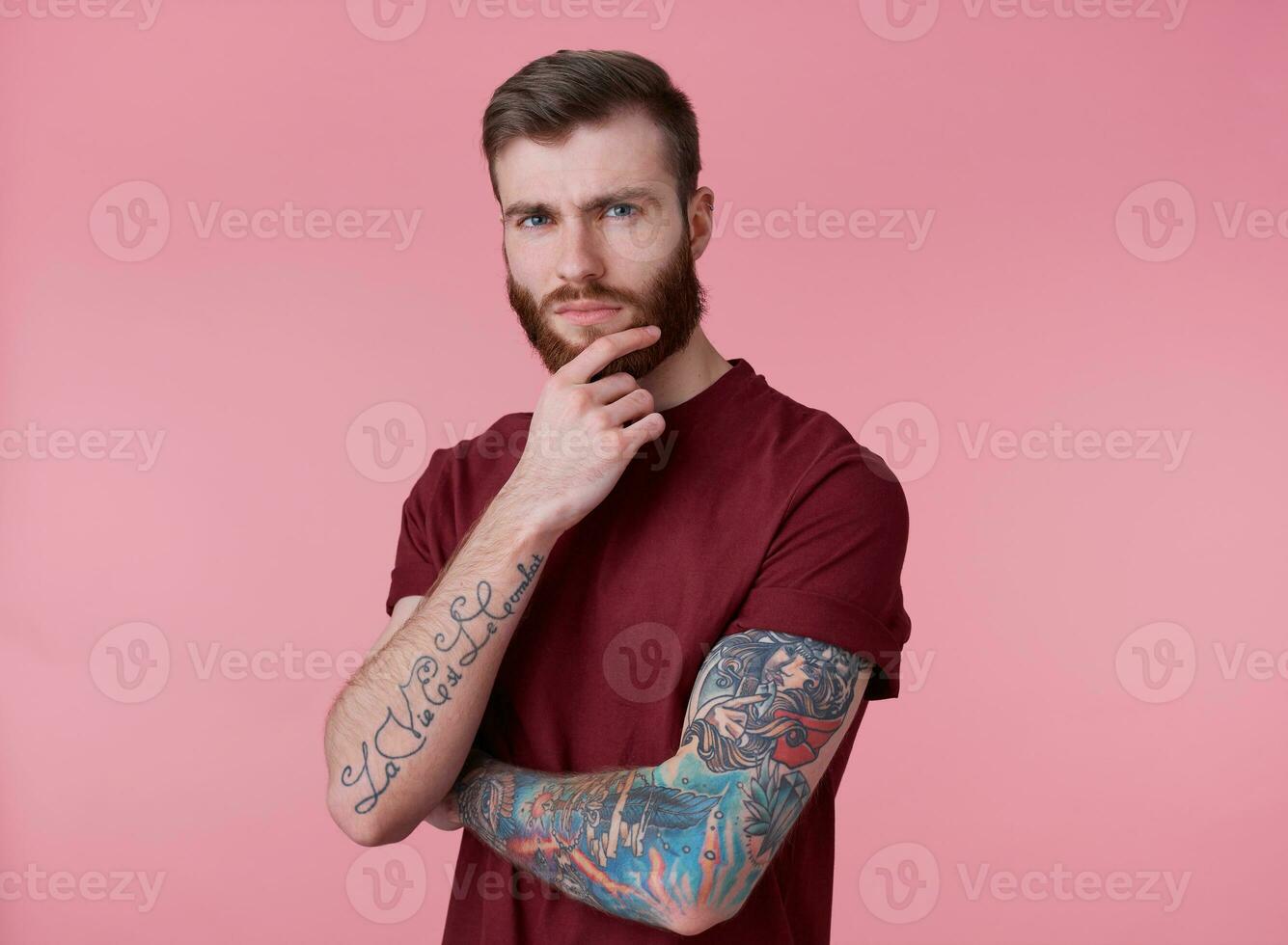 Foto von jung attraktiv Denken tätowiert rot bärtig Mann im rot T-Shirt, steht Über Rosa Hintergrund, sieht aus beim das Kamera und berührt das kinn.