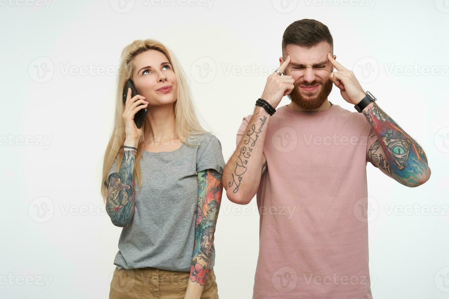 betont jung attraktiv bärtig Kerl runzelte die Stirn seine Gesicht mit geschlossen Augen und behalten Zeigefinger auf Tempel während seine positiv blond tätowiert weiblich haben Telefon Konversation foto