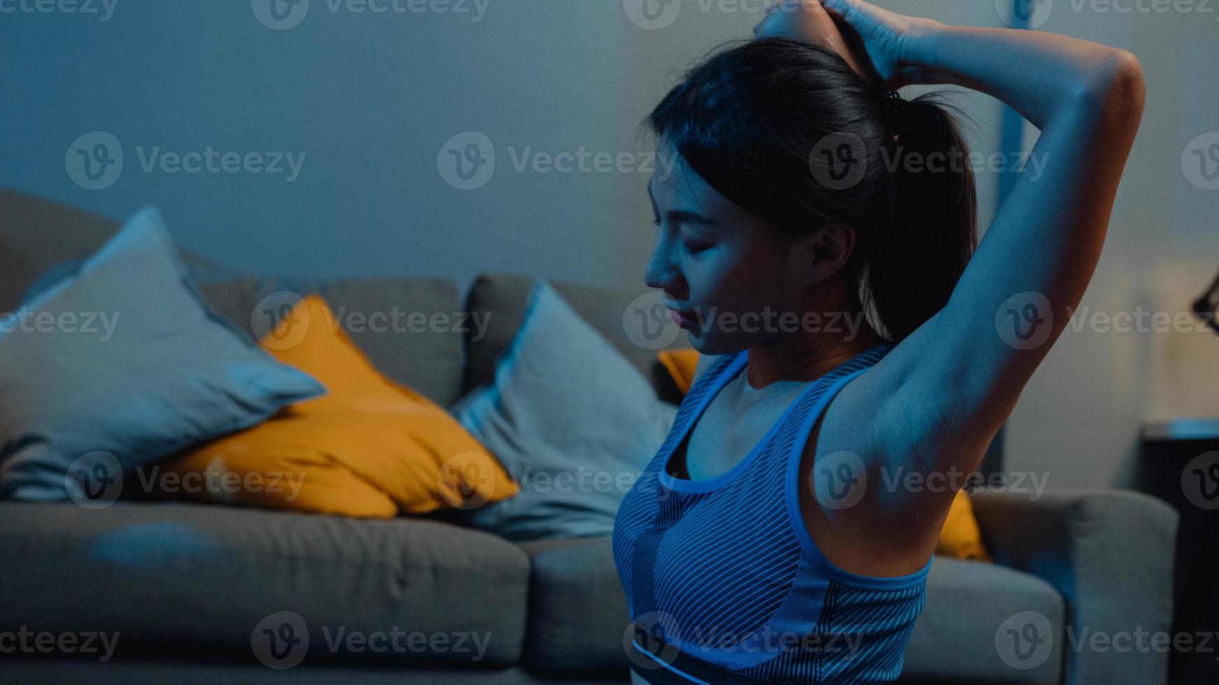 junge asiatische Dame in Sportkleidung macht Yoga-Übungen, die nachts zu Hause im Wohnzimmer trainieren. Sport- und Freizeitaktivitäten, soziale Distanzierung, Quarantäne für das Konzept der Corona-Virus-Prävention. foto