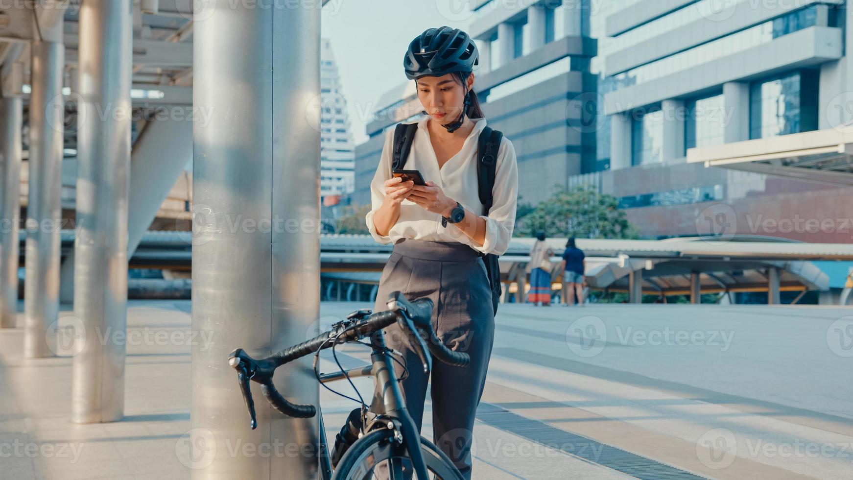 Lächeln Sie asiatische Geschäftsfrau mit Rucksack, die eine Smartphone-Look-Kamera im Stadtstand an der Straße mit dem Fahrrad verwendet, um im Büro zu arbeiten. Sportmädchen benutzen Telefon für die Arbeit. zur Arbeit pendeln, Berufspendler in der Stadt. foto