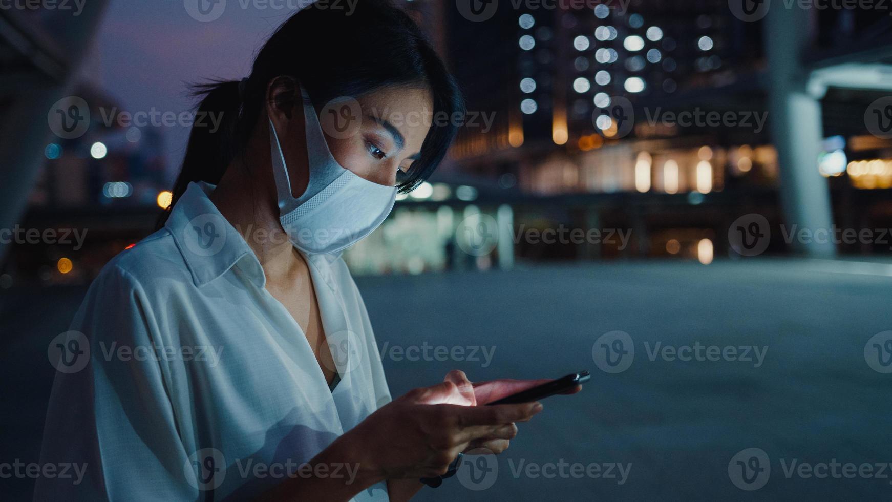 junge asiatische Geschäftsfrau in modischer Kleidung mit Gesichtsmaske mit Smartphone, die Textnachrichten eingibt, während sie nachts in der Stadt im Freien stehen. soziale Distanzierung, um die Ausbreitung des Covid-19-Konzepts zu verhindern. foto