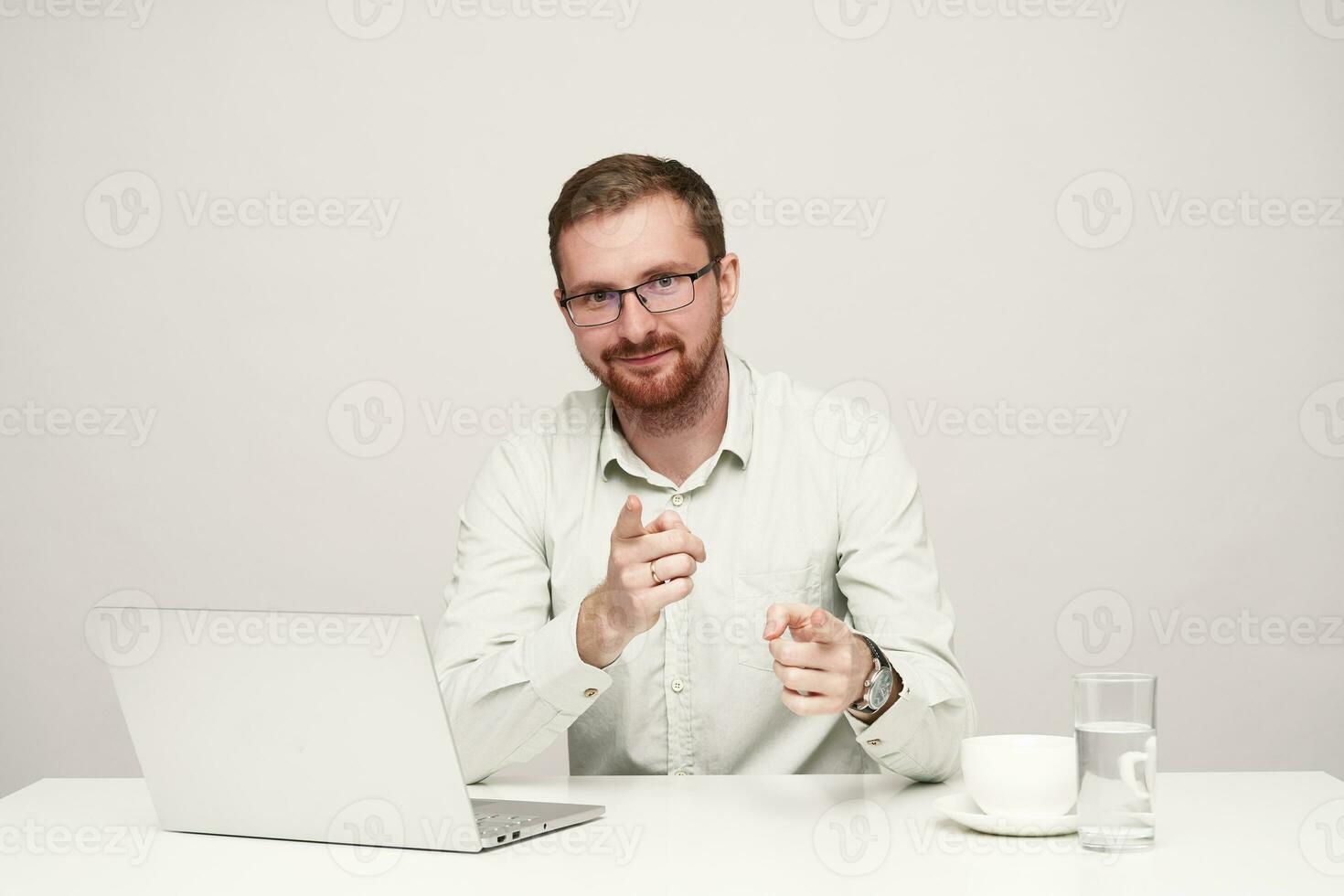 froh jung ziemlich bärtig männlich im Brillen lächelnd positiv während zeigen mit angehoben Zeigefinger beim Kamera, Sitzung beim Tabelle gegen Weiß Hintergrund foto