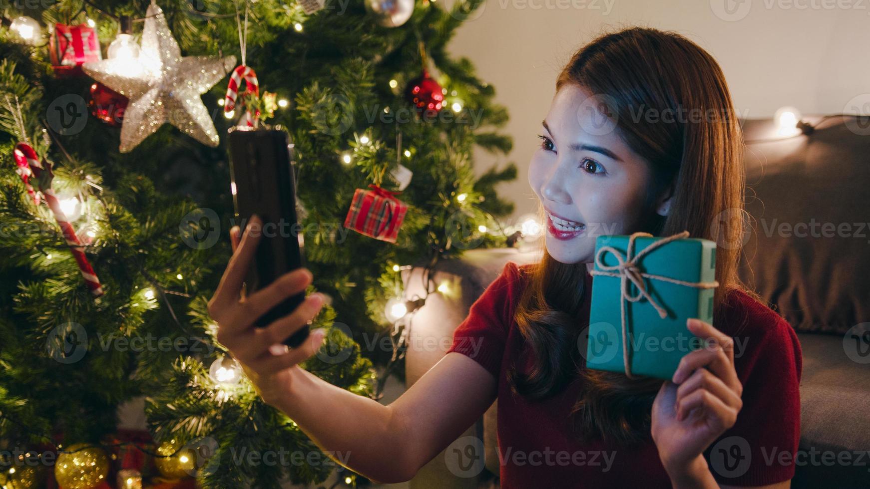 junge asiatische Frau mit Smartphone-Videoanruf im Gespräch mit Paar mit Weihnachtsgeschenkbox, Weihnachtsbaum mit Ornament im Wohnzimmer zu Hause verziert. Weihnachtsnacht und Neujahrsfest. foto