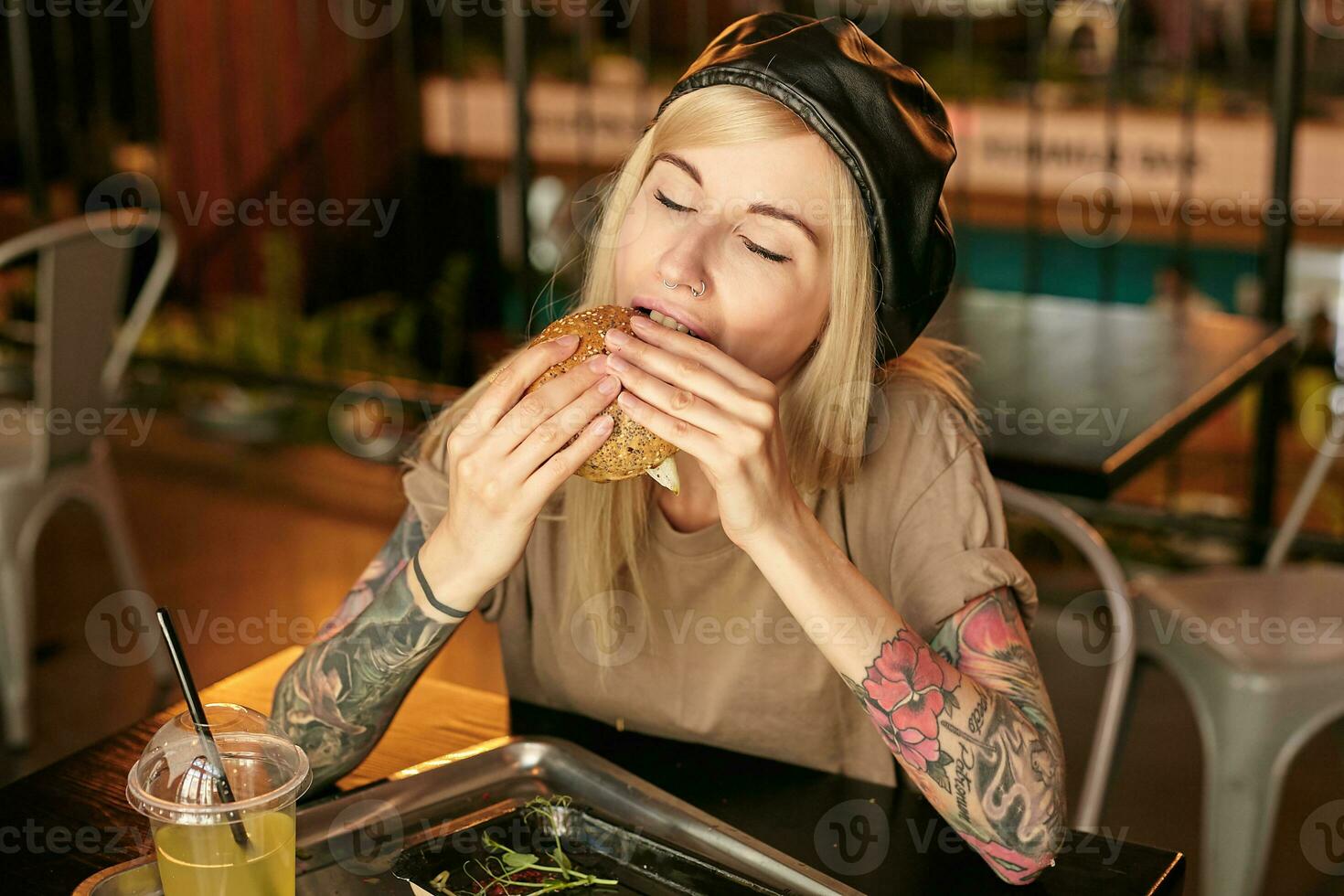 horizontal Foto von ziemlich jung blond Frau mit Tätowierungen posieren Über Stadt Cafe Innere, genießen Geschmack von Hamburger mit geschlossen Augen, tragen modisch Kleider