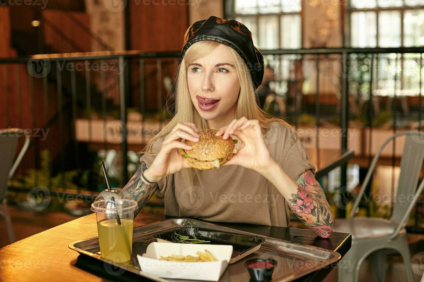 horizontal Schuss von schön jung blond Frau mit Tätowierungen tragen modisch Kleidung, Sitzung beim Tabelle im Stadt Cafe und haben köstlich Mittagessen, suchen beiseite fröhlich und zeigen Zunge foto