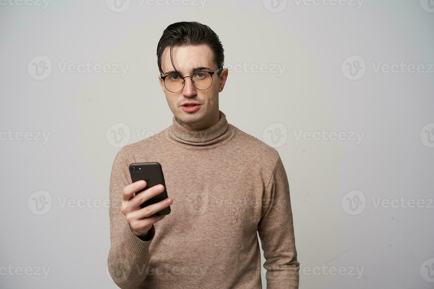 Innen- Schuss von jung dunkel behaart männlich im Brillen suchen beim Kamera während behalten Handy, Mobiltelefon Telefon im angehoben Hand, tragen modisch Kleider während posieren Über Weiß Hintergrund foto