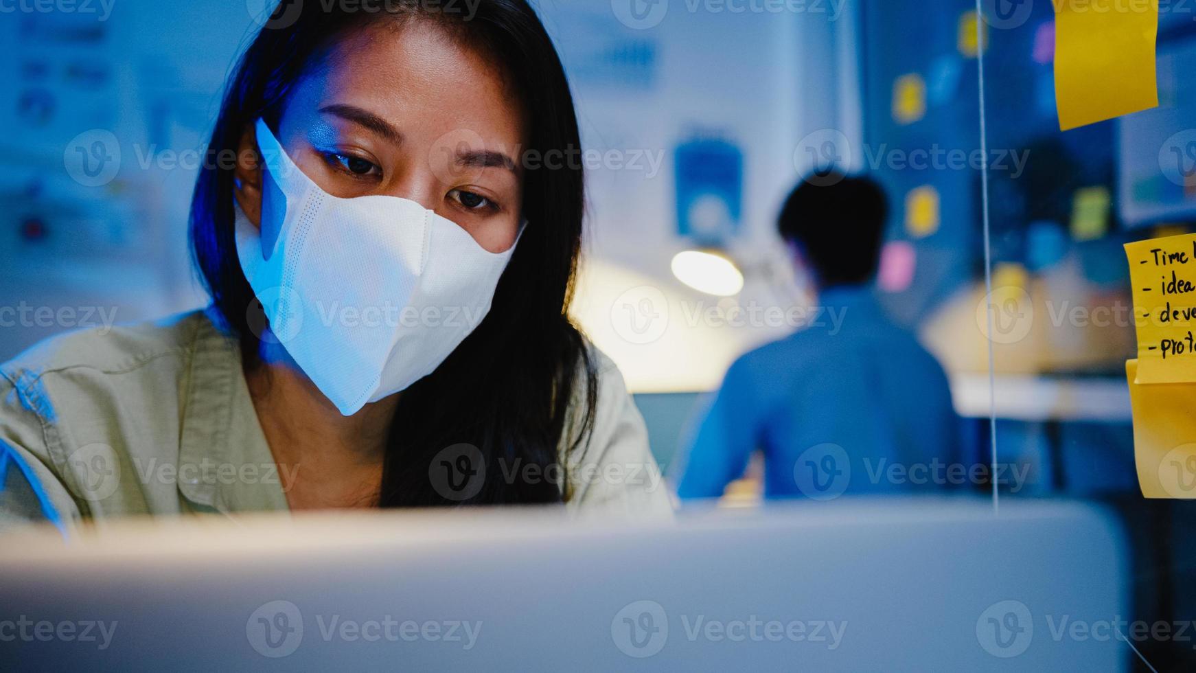 Fröhliche Geschäftsfrau in Asien, die eine medizinische Gesichtsmaske für soziale Distanzierung in einer neuen normalen Situation zur Virenprävention trägt, während sie in der Büronacht den Laptop bei der Arbeit verwendet. Leben und Arbeiten nach dem Coronavirus. foto