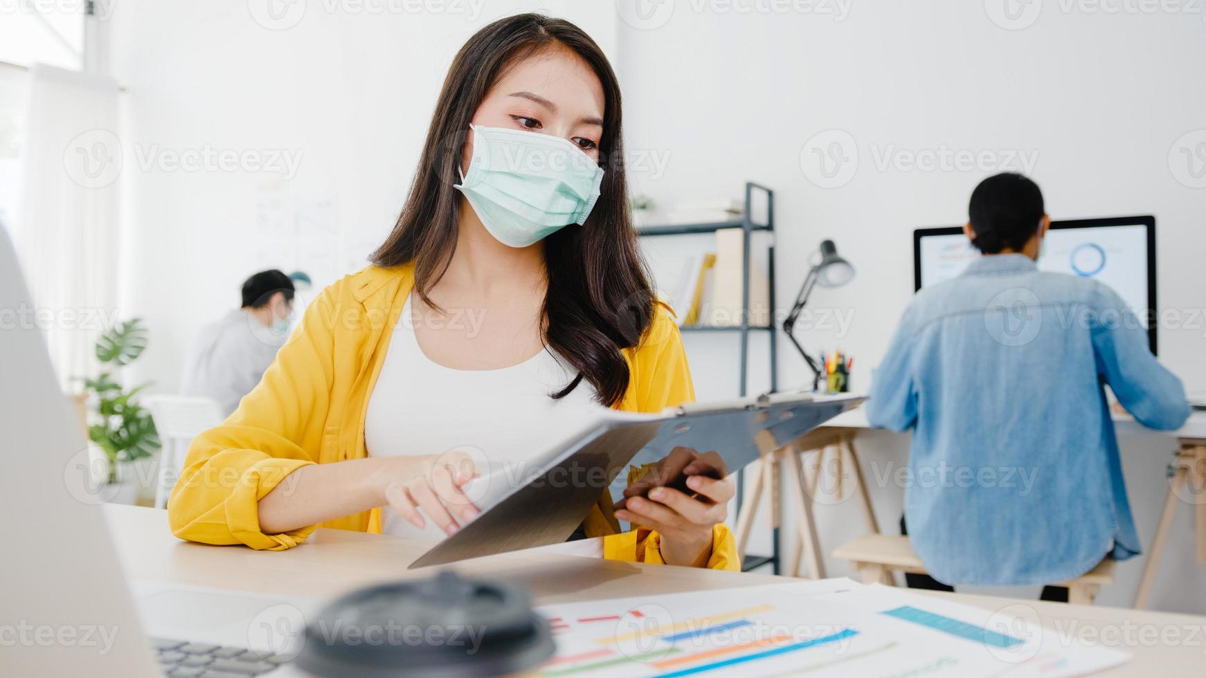 Asiatische Geschäftsfrau, die eine medizinische Gesichtsmaske für soziale Distanzierung in einer neuen normalen Situation zur Virusprävention trägt, während sie den Laptop wieder bei der Arbeit im Büro verwendet. Lebensstil nach dem Corona-Virus. foto