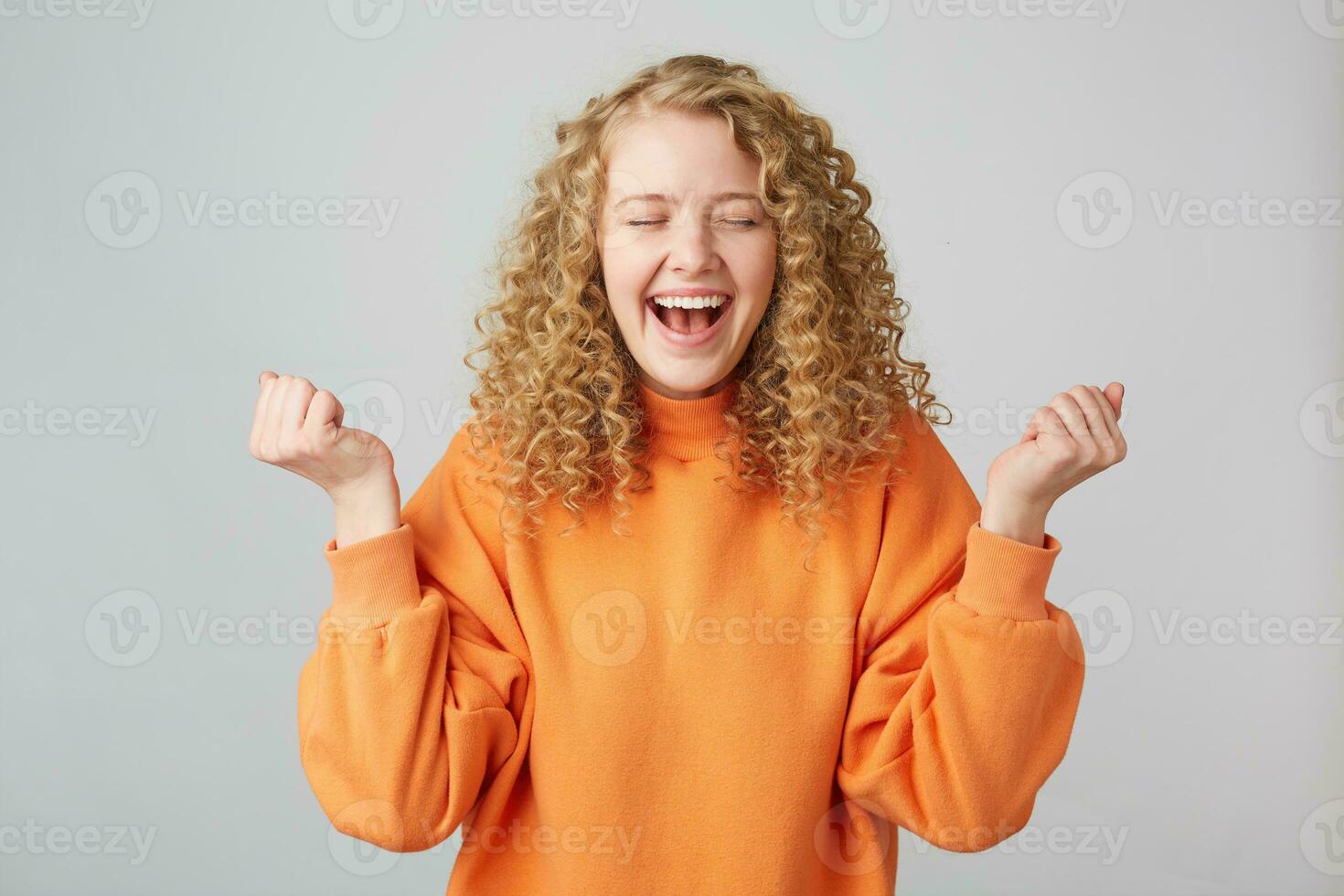 horizontal Porträt von ein heiter glücklich blond Frau im Orange Sweatshirt lächelnd und Zusammenpressen Fäuste mögen Gewinner mit Augen geschlossen im Vergnügen, isoliert Über Weiß Hintergrund foto