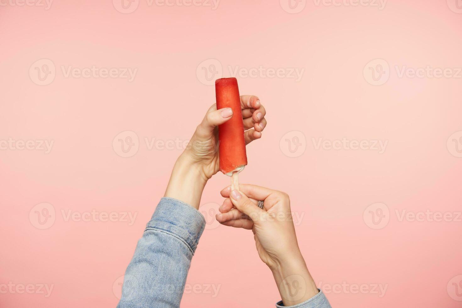 horizontal Schuss von jung weiblich Hände mit nackt Maniküre posieren Über Rosa Hintergrund mit Eis auf Stock mit rot Glasur. Essen Fotografie Konzept foto