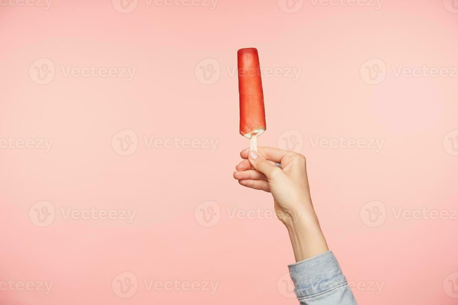 horizontal Foto von jung Frau Hand halten Eis auf Stock mit rot Glasur, isoliert Über Rosa Hintergrund. Essen Fotografie und Mensch Hände Konzept