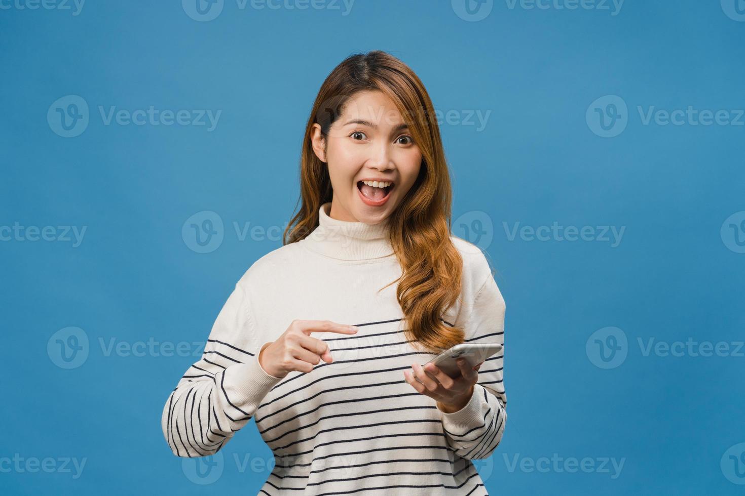 überraschte junge asiatische dame, die handy mit positivem ausdruck benutzt, breit lächelt, in lässiger kleidung gekleidet ist und die kamera auf blauem hintergrund betrachtet. glückliche entzückende frohe frau freut sich über erfolg. foto