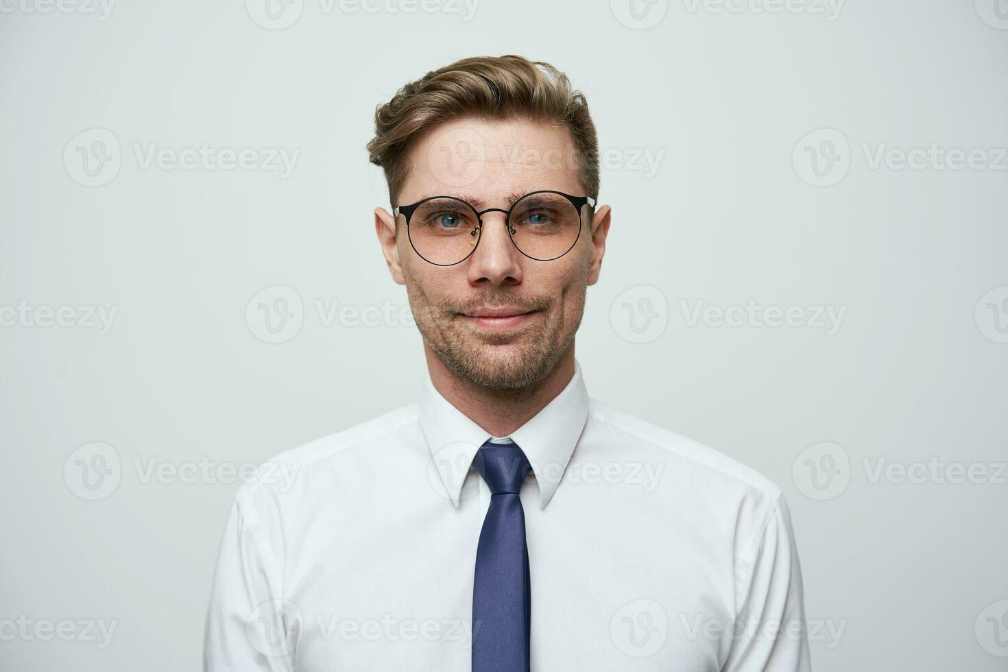 Foto wie auf Reisepass. jung Mann mit stilvoll Haarschnitt, Brille, blau Augen aussehen Gerade wenig lächelnd und selbst gesammelt, gekleidet im Weiß Hemd und Blau binden, unrasiert, Über Weiß Hintergrund
