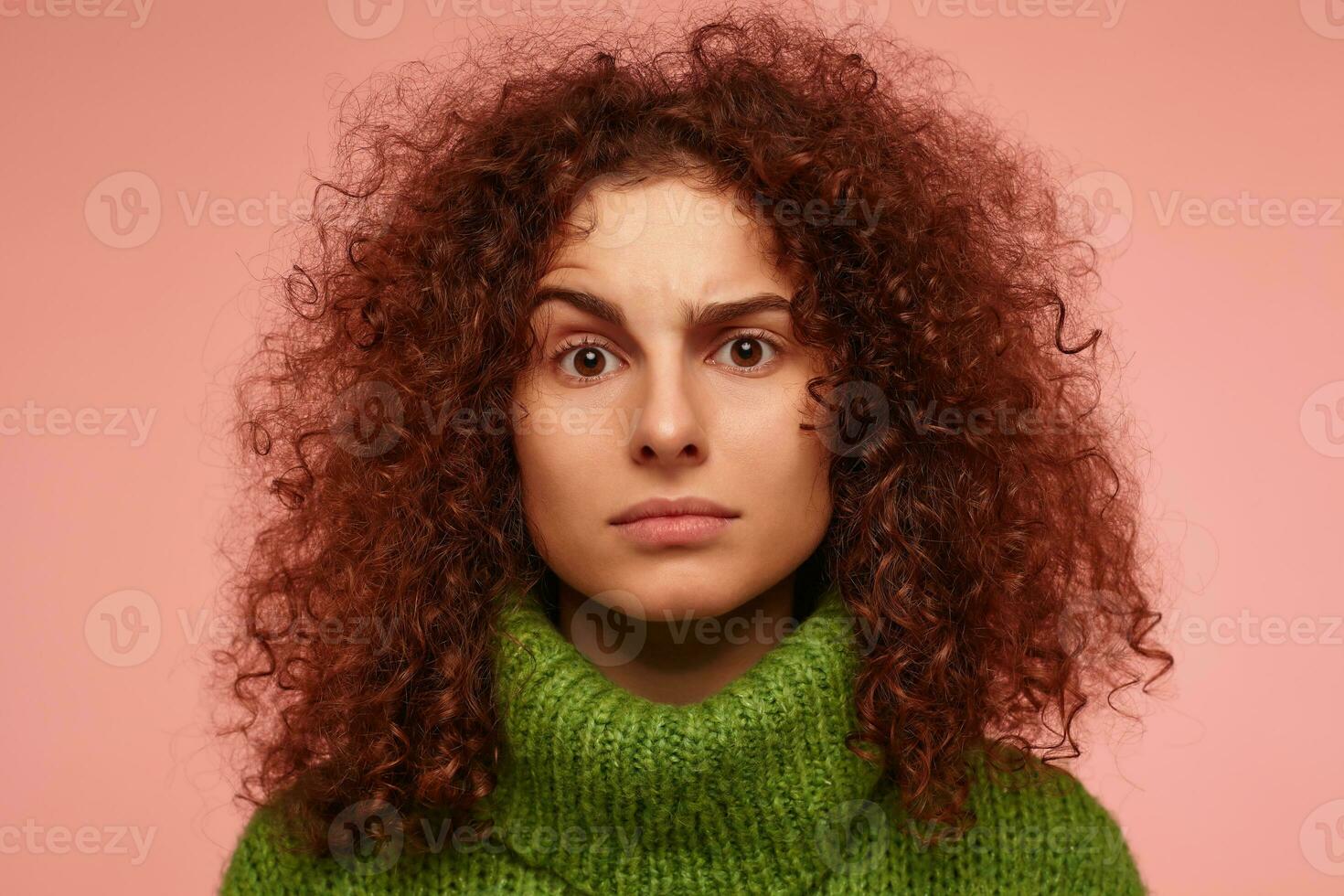 Teenager Mädchen, ernst suchen Rothaarige Frau mit lockig Haar. tragen Grün Rollkragen Sweatshirt und Aufzüge oben ein Augenbraue. Aufpassen beim das Kamera isoliert, Nahansicht Über Pastell- Rosa Hintergrund foto