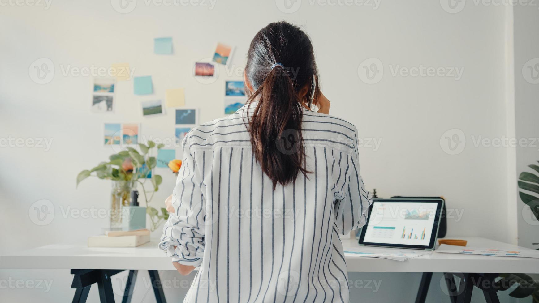 Junge Asia Business Lady Anruf Handy mit Kollegen im Unternehmen über Arbeitsfinanzierung Diagramm Marktplan in Laptop und Tablet im Haus. Mädchen lernen online, arbeiten von zu Hause aus. foto