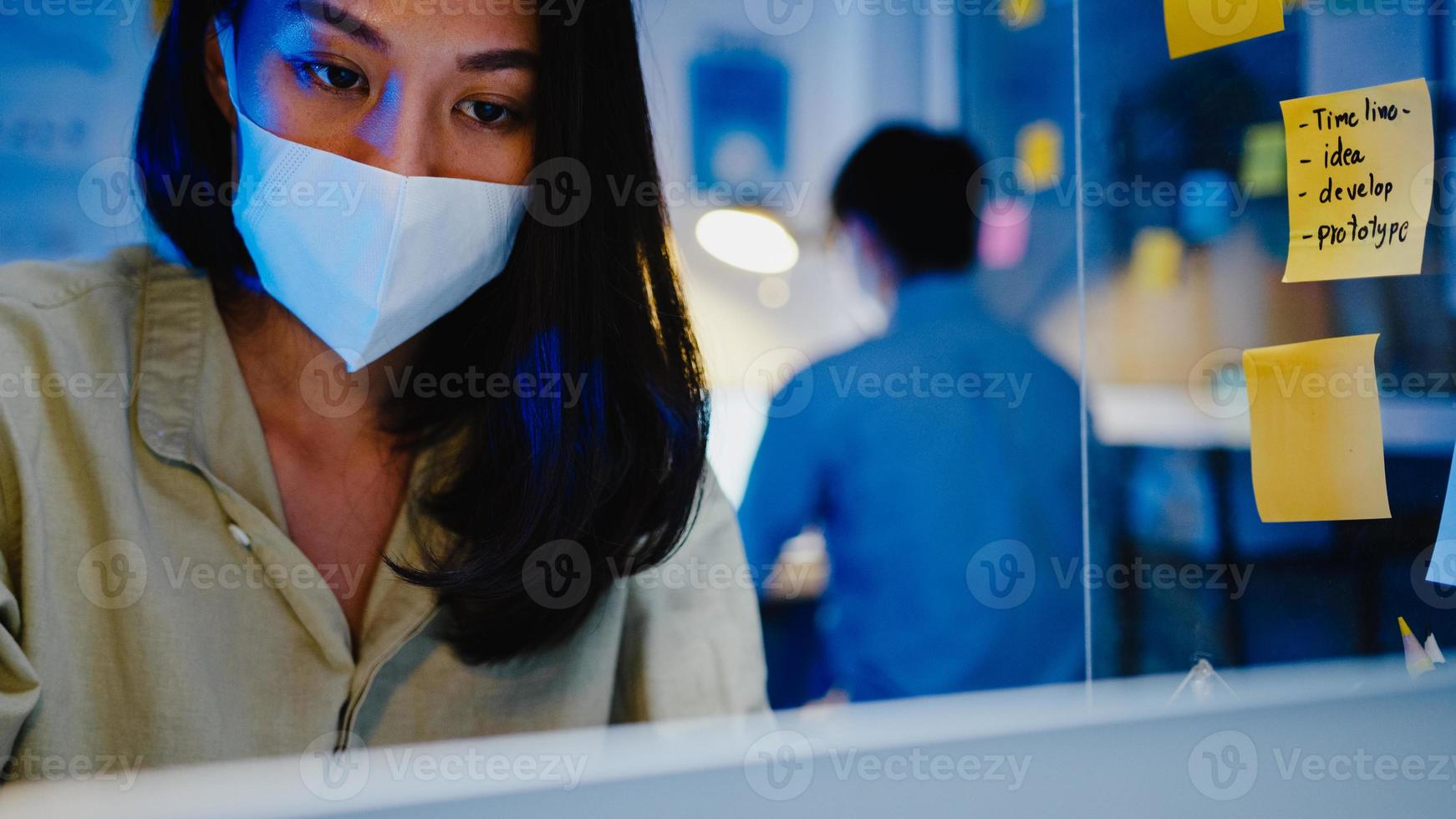 Fröhliche Geschäftsfrau in Asien, die eine medizinische Gesichtsmaske für soziale Distanzierung in einer neuen normalen Situation zur Virenprävention trägt, während sie in der Büronacht den Laptop bei der Arbeit verwendet. Leben und Arbeiten nach dem Coronavirus. foto