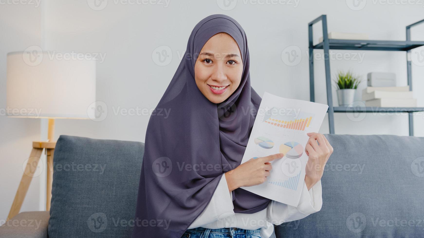 asiatische muslimische dame tragen hijab verwenden computer-laptop sprechen mit kollegen über verkaufsbericht in videoanrufbesprechung, während sie von zu hause aus im wohnzimmer arbeiten. soziale Distanzierung, Quarantäne wegen Corona-Virus. foto