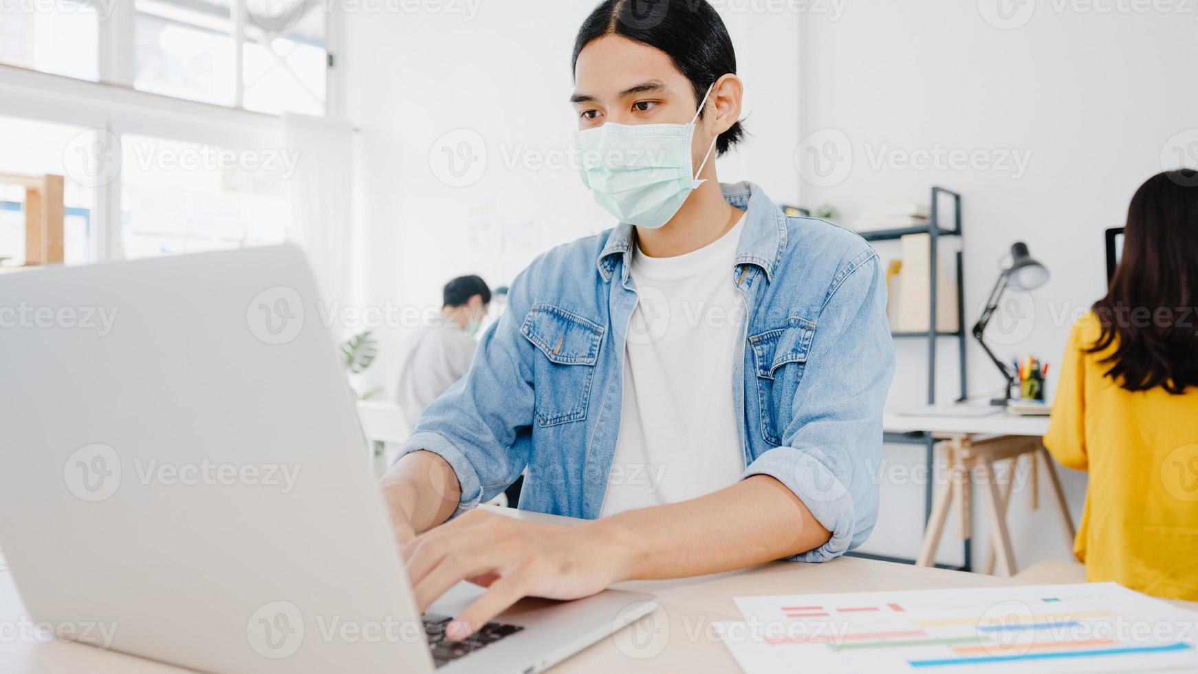 Asiatischer Geschäftsmann, der eine medizinische Gesichtsmaske für soziale Distanzierung in einer neuen normalen Situation zur Virusprävention trägt, während er den Laptop wieder bei der Arbeit im Büro verwendet. Lebensstil nach dem Corona-Virus. foto