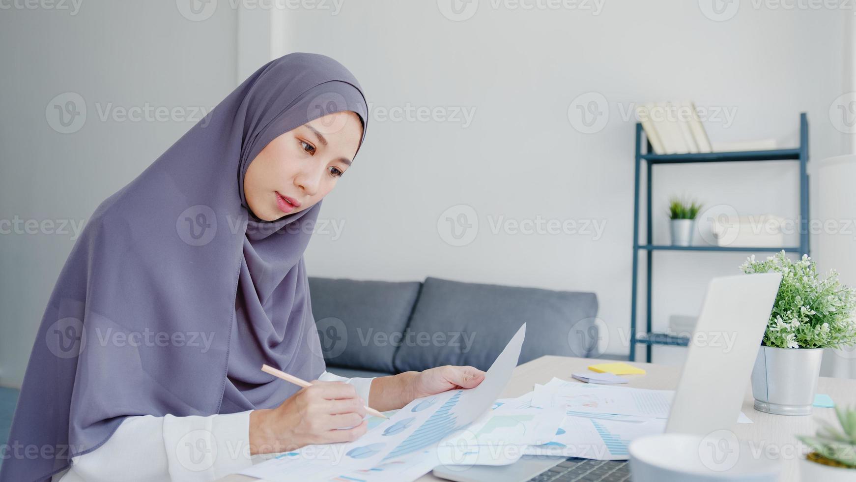 Schöne asiatische muslimische Dame in Kopftuch-Freizeitkleidung mit Laptop im Wohnzimmer im Haus. Fernarbeit von zu Hause aus, neuer normaler Lebensstil, soziale Distanzierung, Quarantäne zur Vorbeugung von Coronaviren. foto