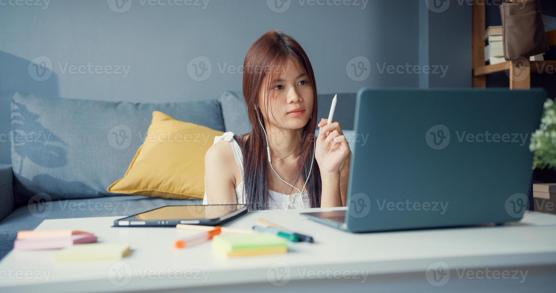 junge asiatische Mädchen Teenager mit lässigem Hemd tragen Kopfhörer mit Laptop lernen online Vorlesung im Laptop im Wohnzimmer im Haus schreiben. Isolieren Sie das Online-E-Learning-Konzept für die Coronavirus-Pandemie. foto