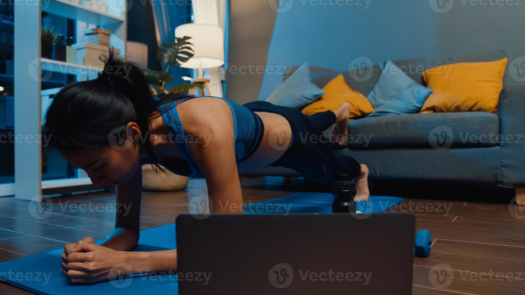 junge asiatische Dame in Sportbekleidung trainiert und benutzt Laptop, um Yoga-Video-Tutorials in der Nacht zu Hause zu sehen. Ferntraining mit Personal Trainer, soziale Distanz, Online-Bildungskonzept. foto
