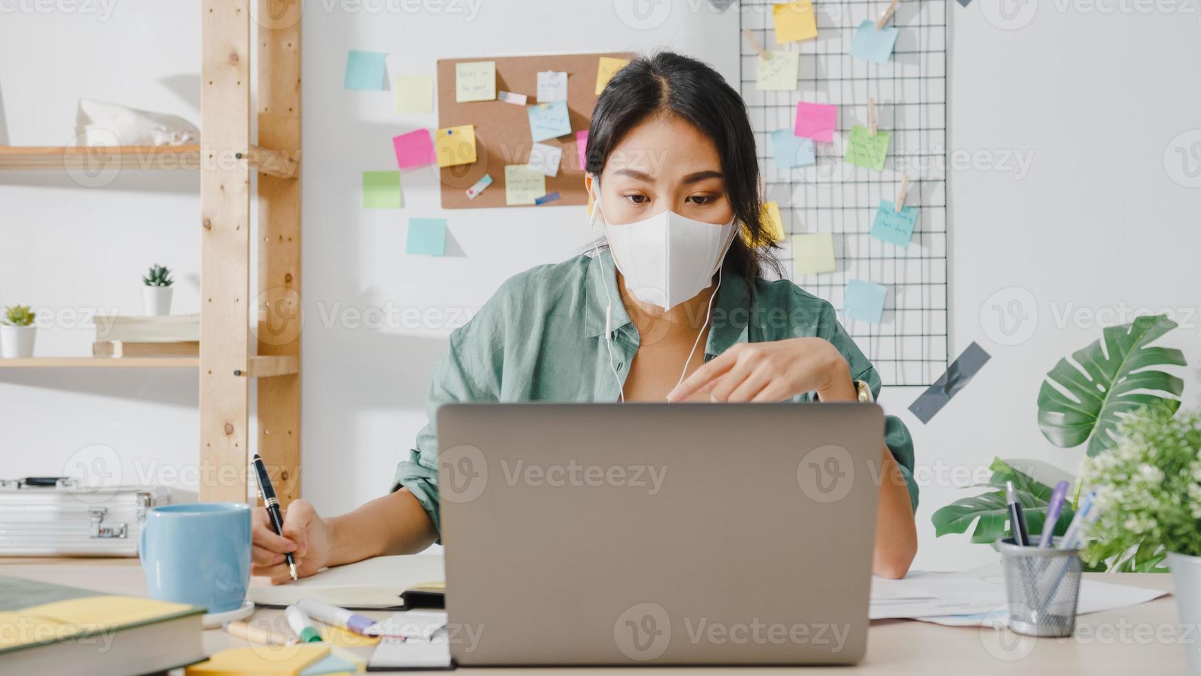 Asiatische Geschäftsfrau, die eine medizinische Gesichtsmaske mit Laptop trägt, spricht mit Kollegen über den Plan im Videoanruf, während sie von zu Hause aus im Wohnzimmer arbeitet. soziale Distanzierung, Quarantäne zur Vorbeugung des Coronavirus. foto